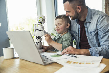 Smiling man looking at son playing with robot at home - UUF23389