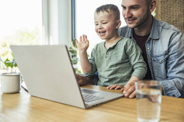 Glücklicher Junge winkt mit der Hand, während er mit seinem Vater zusammensitzt und einen Videoanruf über einen Laptop zu Hause tätigt - UUF23391