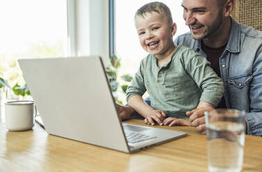 Fröhlicher Sohn und Vater mit Laptop sitzend bei einem Videoanruf zu Hause - UUF23392