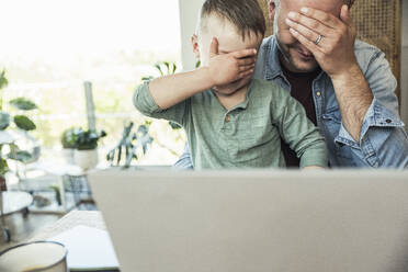 Father and son covering eyes with hands while sitting with laptop at home - UUF23393