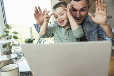 Mischievous father and son teasing while sitting with laptop at home - UUF23394