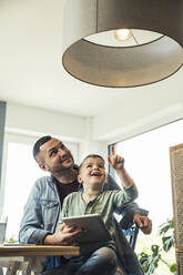 Smiling boy pointing at pendant light while sitting with father in smart home - UUF23400