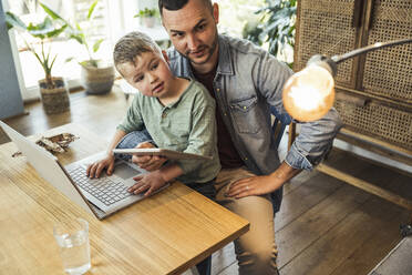 Vater und Sohn betrachten eine Glühbirne, während sie mit Tablet und Laptop in einem intelligenten Haus sitzen - UUF23401