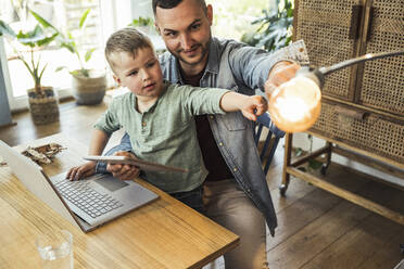 Mann und Sohn zeigen auf Glühbirne, während sie mit Tablet und Laptop im intelligenten Haus sitzen - UUF23402