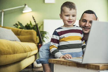 Lächelnder Vater mit süßem Sohn, der im Wohnzimmer zu Hause ein Video auf dem Laptop ansieht - UUF23404