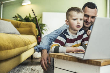 Lächelnder Vater sieht seinen Sohn an, der im Wohnzimmer einen Laptop benutzt - UUF23405