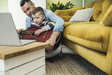 Junge, der Gitarre spielen lernt, sitzt mit seinem Vater im Wohnzimmer und sieht sich ein Online-Video auf seinem Laptop an - UUF23407