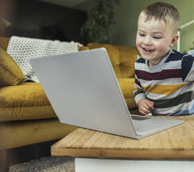 Smiling cute boy watching videos on laptop in living room at home - UUF23408