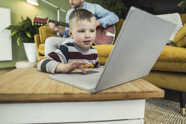 Cute boy using laptop while father playing guitar in living room - UUF23409