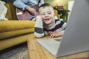 Cute boy watching video on laptop while father playing guitar in living room - UUF23410
