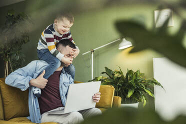 Man using laptop while mischievous son sitting on shoulder in living room - UUF23411