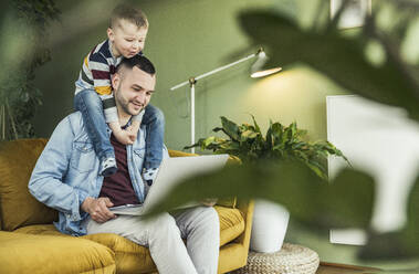 Smiling man using laptop while son sitting on shoulder in living room at home - UUF23412