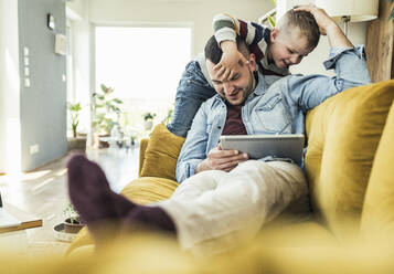 Schelmischer Sohn spielt mit Vater mit Tablet auf Sofa im Wohnzimmer sitzend - UUF23414