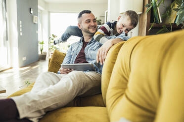 Smiling man sitting with tablet looking at son playing in living room at home - UUF23415