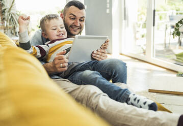 Smiling father and son watching video through tablet while sitting on sofa in living room - UUF23416