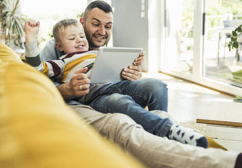 Lächelnder Vater und Sohn sehen sich ein Video über ein Tablet an, während sie auf dem Sofa im Wohnzimmer sitzen - UUF23416
