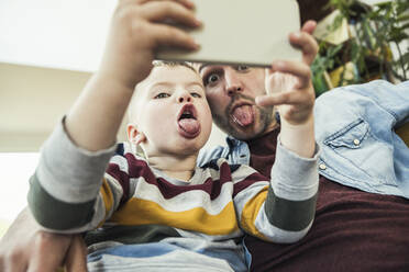 Father sticking out tongue with son talking selfie through smart phone at home - UUF23418