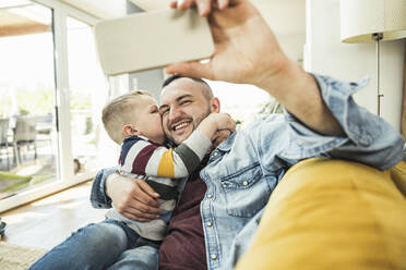 Son kissing on cheek while father taking selfie selfie through mobile phone on living room - UUF23419