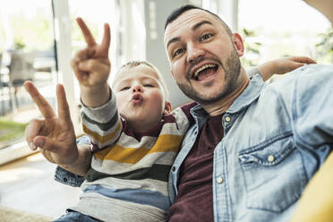 Happy father and son gesturing peace sign in living room at home - UUF23420