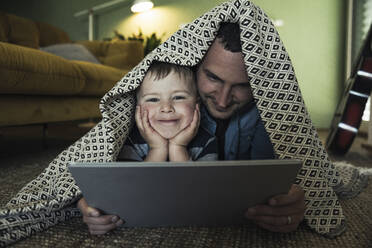 Lächelnder Vater und Sohn mit digitalem Tablet unter einer Decke im Wohnzimmer liegend - UUF23421