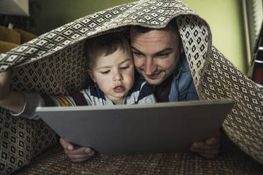 Father and son watching video while lying under blanket in living room at home - UUF23422