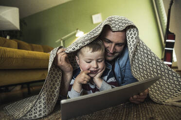 Happy father and son watching video while lying under blanket in living room at home - UUF23423