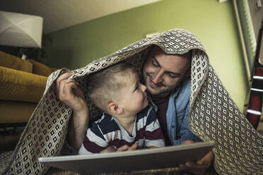 Smiling father and son looking at each other while under blanket in living room at home - UUF23424