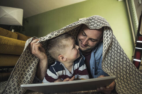 Lächelnder Vater und Sohn, die sich unter einer Decke im Wohnzimmer zu Hause ansehen - UUF23424