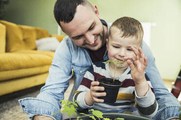 Smiling father sitting with son holding potted plants in living room - UUF23425