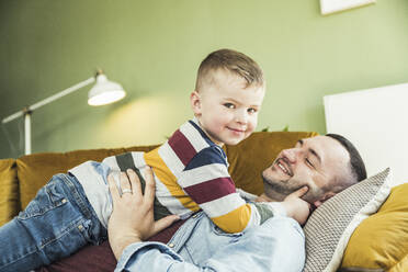 Cute boy lying with father on sofa in living room at home - UUF23427