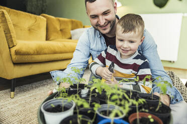 Cute boy spraying water on potted plants while sitting with father in living room at home - UUF23429
