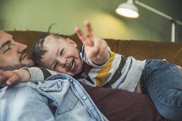 Cheerful boy gesturing peace sign while lying with father sleeping in living room - UUF23430
