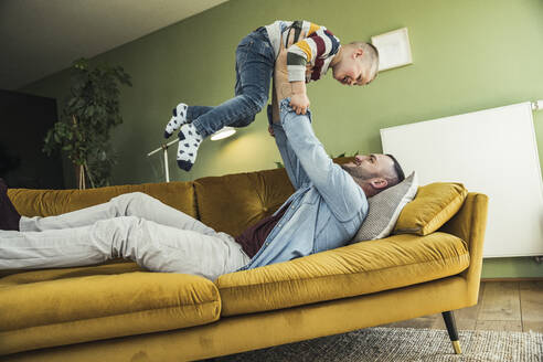 Verspielter Vater hebt seinen Sohn hoch, während er auf dem Sofa im Wohnzimmer liegt - UUF23432
