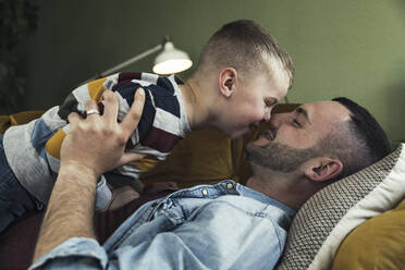 Happy father embracing son while lying on sofa in living room at home - UUF23433