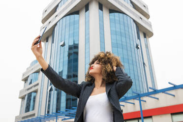 Weibliche Fachkraft mit Hand im Haar, die ein Selfie mit ihrem Handy vor einem Bürogebäude macht - JRVF00631