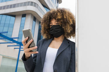 Afro-Geschäftsfrau, die während einer Pandemie ihr Smartphone benutzt und sich an die Wand lehnt - JRVF00634