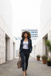 Gut gekleidete Geschäftsfrau mit Handtasche auf dem Fußweg inmitten eines modernen Bürogebäudes - JRVF00638