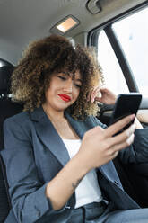 Berufstätige Frau mit lockigem Haar, die im Auto sitzend ein Mobiltelefon benutzt - JRVF00646