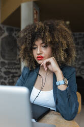 Afro businesswoman wearing headphones sitting in front of laptop at cafe - JRVF00652