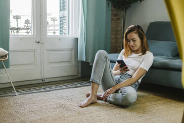 Relaxed woman using mobile phone while sitting on carpet at home - XLGF01838