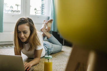 Smiling woman using laptop while lying on carpet at home - XLGF01841