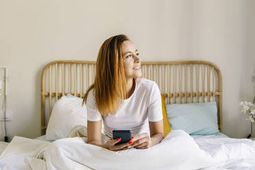 Relaxed woman looking away while holding mobile phone on bed at home - XLGF01882