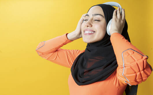 Lächelnde Frau mit geschlossenen Augen hört Musik über drahtlose Kopfhörer vor gelbem Hintergrund - JCCMF02575