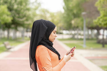 Arabische Frau mit Kopftuch benutzt ein Smartphone in einem öffentlichen Park - JCCMF02576