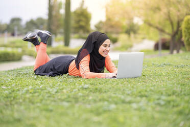 Smiling woman using laptop while lying down in park - JCCMF02590