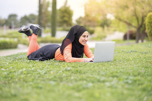 Lächelnde Frau, die einen Laptop benutzt, während sie im Park liegt - JCCMF02590