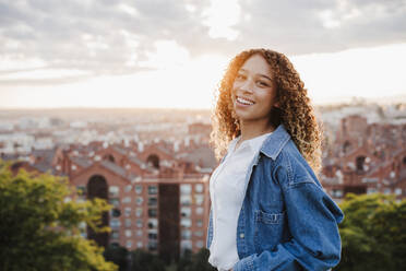 Lächelnde Frau in Jeansjacke in der Stadt bei Sonnenuntergang - EBBF03641