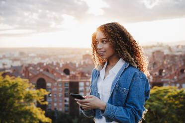 Hispanische Frau schaut weg, während sie bei Sonnenuntergang ihr Smartphone hält - EBBF03646