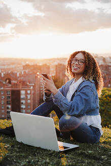Lächelnde Frau mit Smartphone und Laptop im Park sitzend bei Sonnenuntergang - EBBF03655