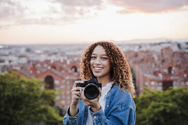 Lächelnde Frau hält Kamera bei Sonnenuntergang - EBBF03663
